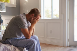 Person with short hair who is wearing pajamas sits on end of bed with head in hands, pose of worry