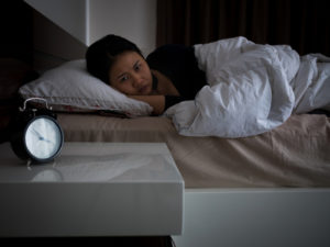 Adult person of color with hair pulled back lies in bed in dark, awake, looking at clock in frustration