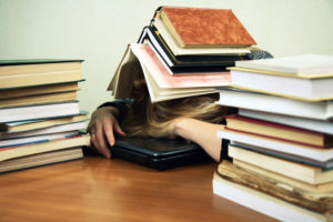 A young woman is sleeping on a laptop with a heap of books on her head.