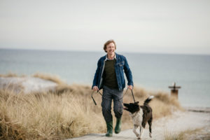 Person with short hair wearing rain boots takes walk along beach with large dog