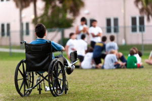 A 12-year-old child sitting in a wheel chair watching other kids getting together in the park while he is left behind.