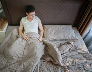Young Asian adult with short hair and glasses sits up in bed under duvet cover looking downcast