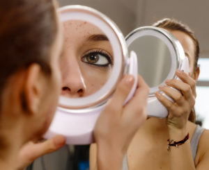 A teenage girl examines her skin in a hand mirror.