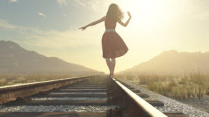 A girl balances on the rail of the train tracks. There are hills in the distance.