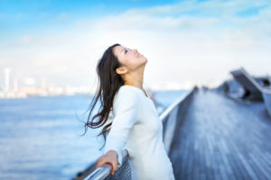 A young woman leans against a railing, smiling at the sky. One can see an out-of-focus ocean and pier behind her.