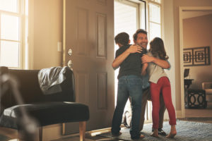 Photo of parent with short hair and facial hair standing in open doorway, hugging two children