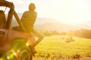 Rear view photo of young adult with short hair sitting on back of Jeep looking out at mountains and field