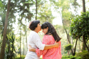 Latin mother kissing daughter with long hair on forehead in sunny, tree-lit area