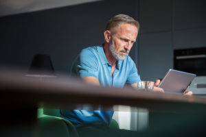 Stressed-looking mature adult with gray hair and short beard looks serious at table while reading on tablet