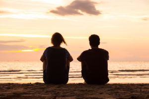 Rear view photo of two friends sitting together on beach at sunset