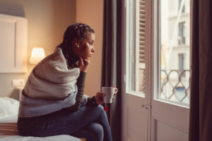 Person sits on edge of bed looking out window, wrapped in blanket, holding tea