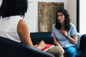 Person in therapy with dark curly hair shares something with hand on chest while therapist listens