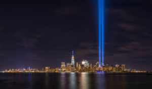 Photo taken from New Jersey shows view of New York City across water with blue lights representing the Twin Towers