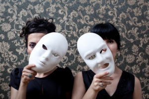 Two people with banks and hair pulled back looking out through masks held to side