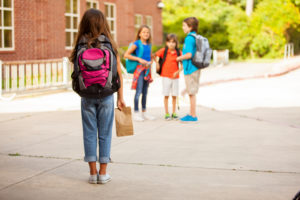A child with a pink backpack and bag lunch stands in schoolyard.