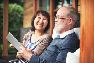 Two older adults sit on bench outside cabin smiling and talking about something they are looking at on a tablet