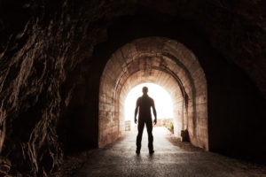 Rear view of person with short hair wearing pants and shirt standing looking out toward bright exit of dark tunnel