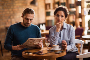 Pensive person with long curly hair sits in a cafe while partner looks down and away, reading newspaper. 