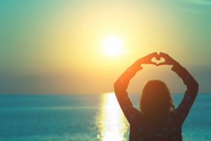 Rear view of person standing by beach looking out to sea with hands forming heart shape