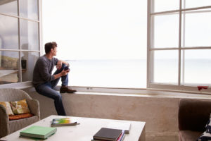 Adult with short hair sits on window seat in spacious room and looks out toward beach