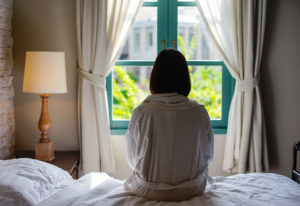 Person with short dark hair sits on bed in robe looking out open window