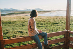 Person with short hair wearing sweater sits on fence and looks into the distance