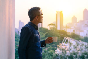 Person in denim shirt holds cup while looking out window of office, thinking