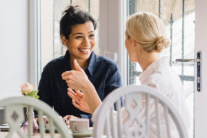 Two adults in their forties, both with hair pulled back, sit at table in cafe, talking happily