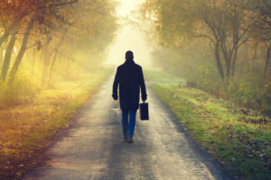 Rear view of a person wearing jeans and coat walking down road with suitcase