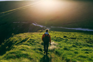 Rear view of young adult in jacket walking along grassy hills with sun above ahead