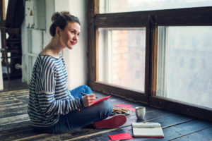 Adult with curly hair pulled back in bun sits on floor, looking out window, working on letter