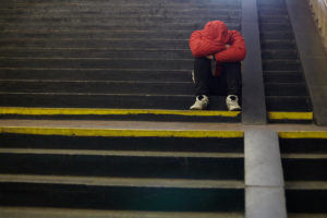 A figure in a red jacket sits on some stairs. Their face and hands are hidden.