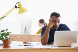 Person with crew cut and some facial hair sits in front of laptop, chin in hand, looking tired and drained