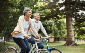 Older couple takes bike ride in tree-lined park. Both are smiling happily