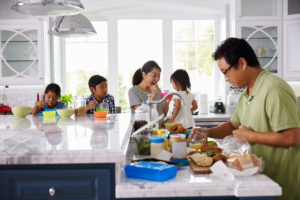 One parent feeds children breakfast while other parent makes lunches in open kitchen