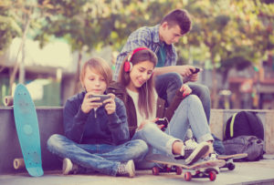 Group of adolescents sitting with skateboards and headphones, all using smartphones