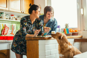 Two women eat eggs at a kitchen counter while a large dog begs for food.