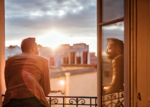 Person with facial hair leans out window to look at city, smiling face shown in reflection