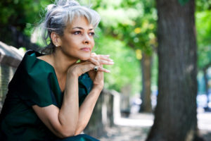 Adult with gray hair pulled back in ponytail sits outside under tree looking seriously into distance