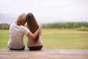 Parent and teen, both with long hair, sit on porch looking out into field. Rear view photo