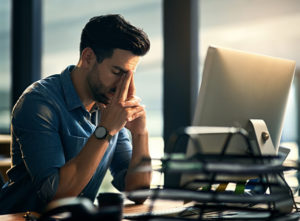 Young business person with short hair and facial hair rubbing eyes with fingertips while resting in front of computer