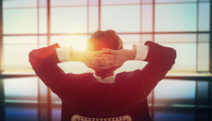 Rear view of person in suit sitting and looking outside large window with hands behind head with sunspot illuminating area