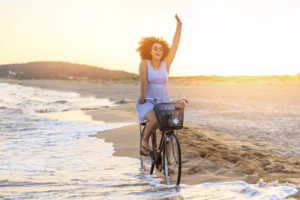 Person in sundress rides bike in waves at beach, waving at someone in distance