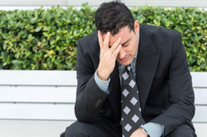 A middle-aged businessman sits on a bench by some shrubbery.