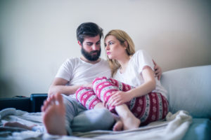 A young man and woman lean into each other as they sit on an unmade bed.