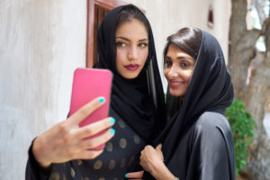 Two young women in hijabs take a selfie with a pink smartphone.