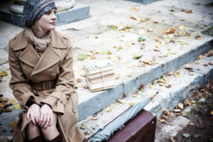 Person with blonde hair wearing hat and coat sits outside looking off into the distance with peaceful expression
