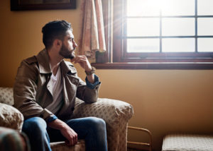 Person with short hair and facial hair sits on sofa with chin in hand looking out window