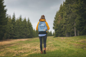 Rear view of person with long hair and backpack walking up hill into field of trees and grass under cloudy sky