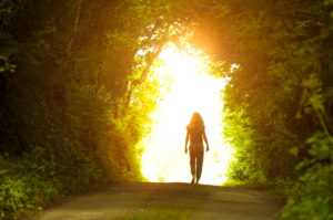 Person with long, shoulder-length hair walks out of wide forest tunnel into afternoon sunlight